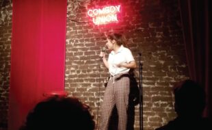 A young woman standing on a stage with a neon sign above her that reads comedy union