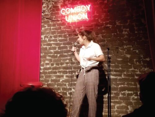 A young woman standing on a stage with a neon sign above her that reads comedy union