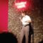 A young woman standing on a stage with a neon sign above her that reads comedy union