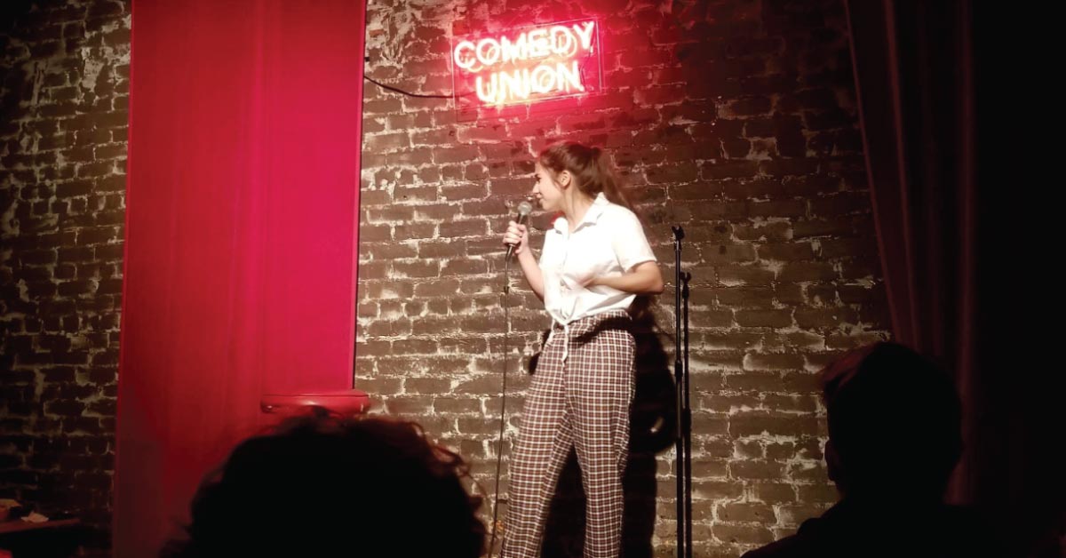 A young woman standing on a stage with a neon sign above her that reads comedy union